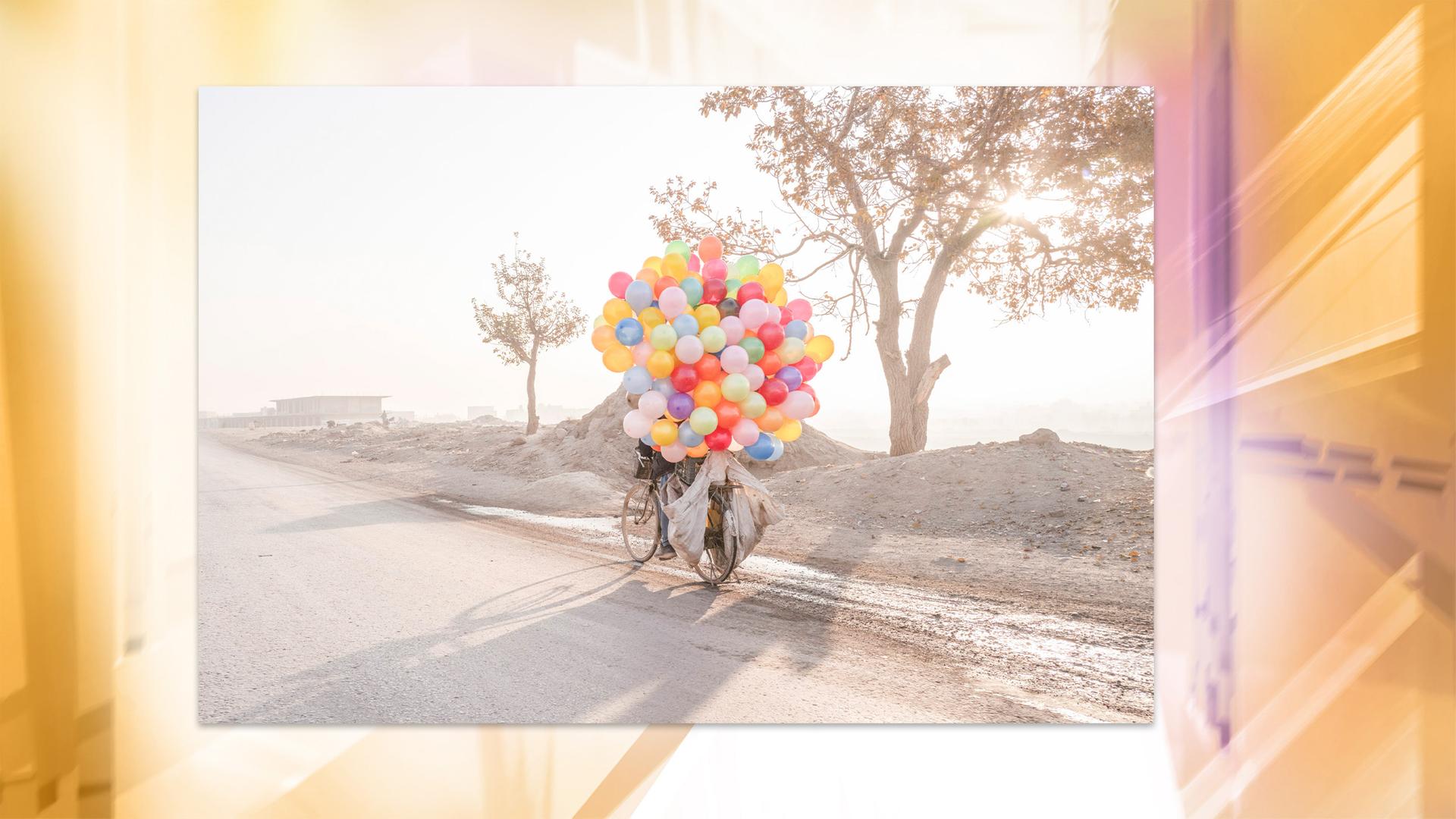 In einer staubigen Landschaft sieht man ein Fahrrad, welches von vielen, bunten Luftballons verdeckt ist.