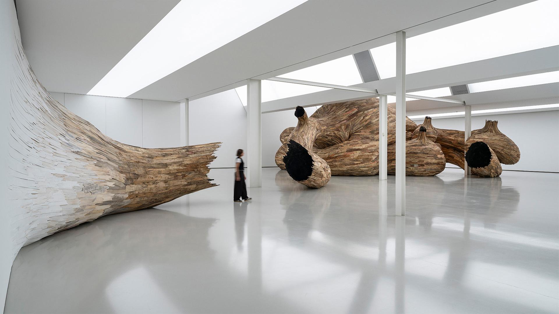 Eine Person steht in einem Ausstellungsraum, in dem die großformatige Installation eines hölzernen Kunstwerkes zu sehen ist.