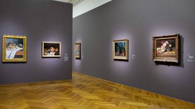 Zwei Wände an denen jeweils mehrere Gemälde hängen. Auf ein paar der Gemälde sind Blumen und Pflanzen zu sehen und auf einem ist eine Obstschale gemalt.