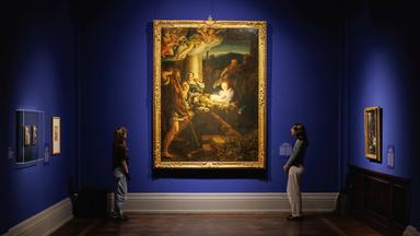 In einem Ausstellungsraum schauen sich zwei Personen ein Gemälde an, welches an einer blauen Wand hängt.