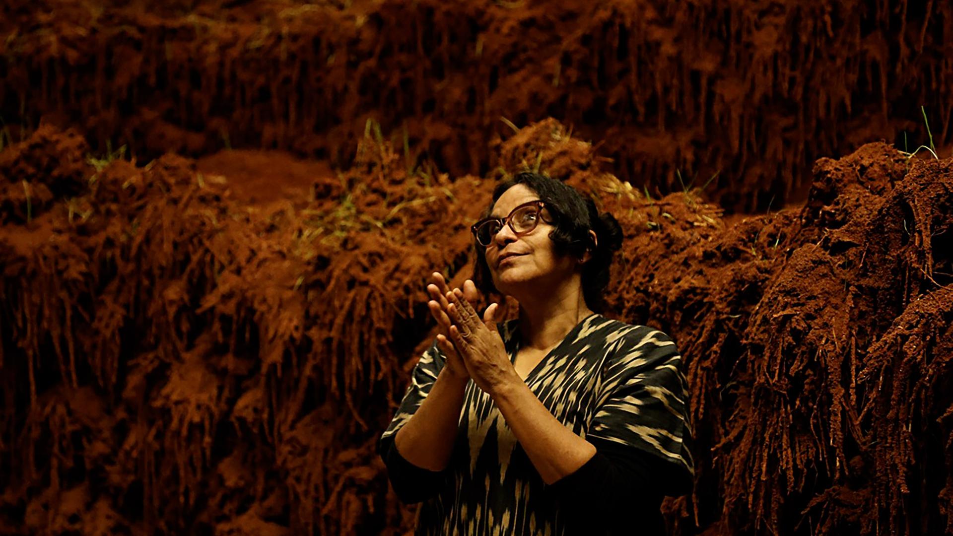 Man sieht eine Frau, die eine Brille trägt, vor einem Kunstwerk aus Erde.