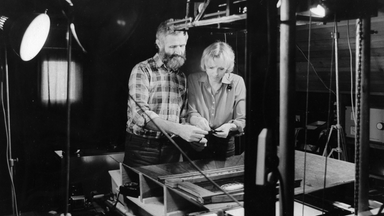 Auf der schwarz-weiß Fotografie sind ein Mann und eine Frau zu sehen, die vor einer Maschine stehen und auf etwas schauen, das sie in ihren Händen halten.