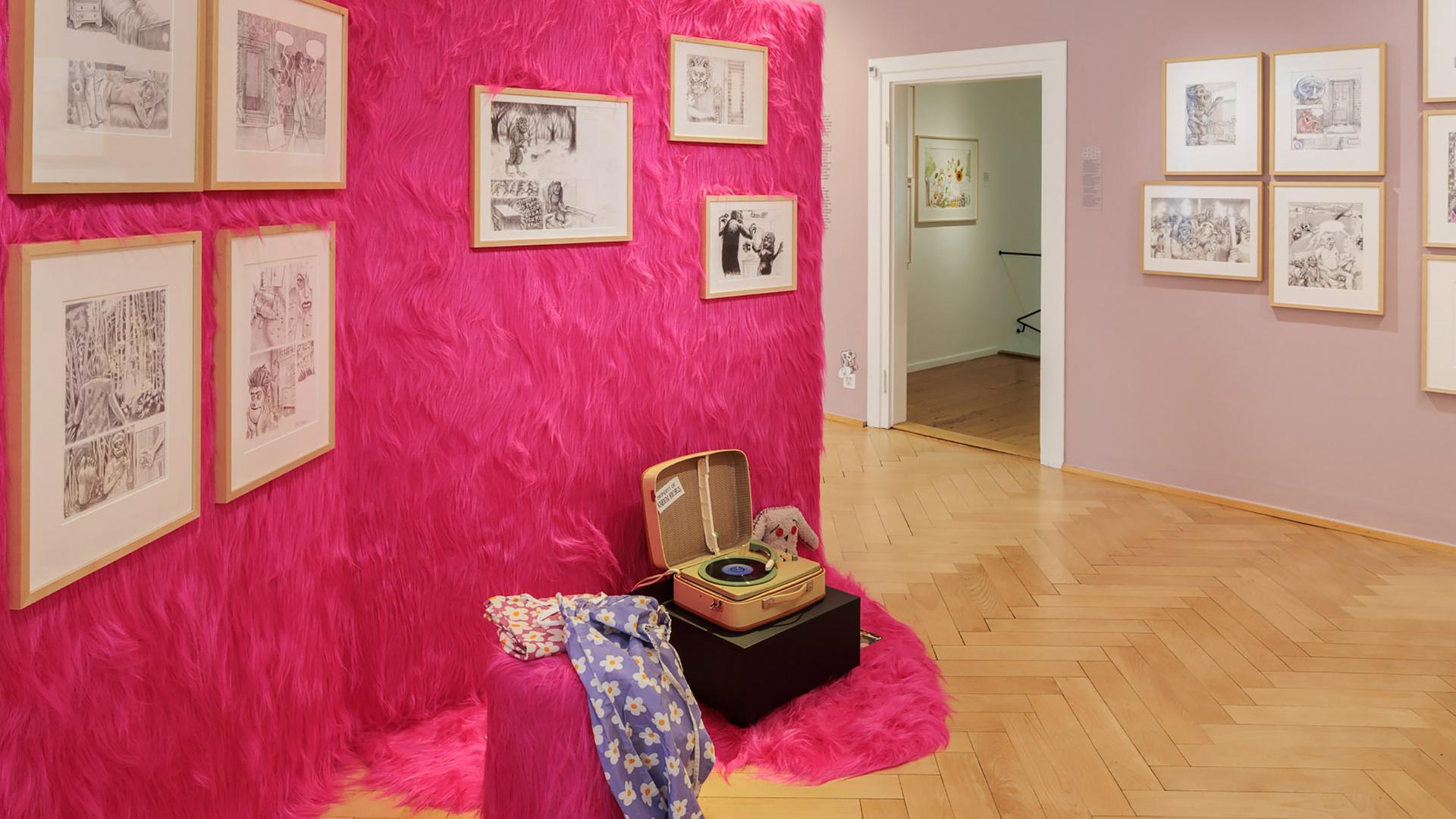 In einem Ausstellungsraum sieht man Illustrationen an einer mit pinkfarbenem Zottelstoff verkleideten Wand hängen. Im Vordergrund steht ein altmodischer Plattenspieler.