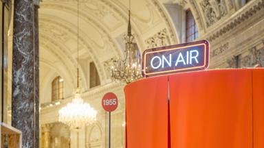 In einem historischen Gebäude mit Marmorsäulen und Kronleuchtern steht in der Mitte ein Schild auf dem "1955" steht, rechts davon sieht man ein Neonleuchtschild "On air", welches sich über einer orangefarbenen Kabine befindet.