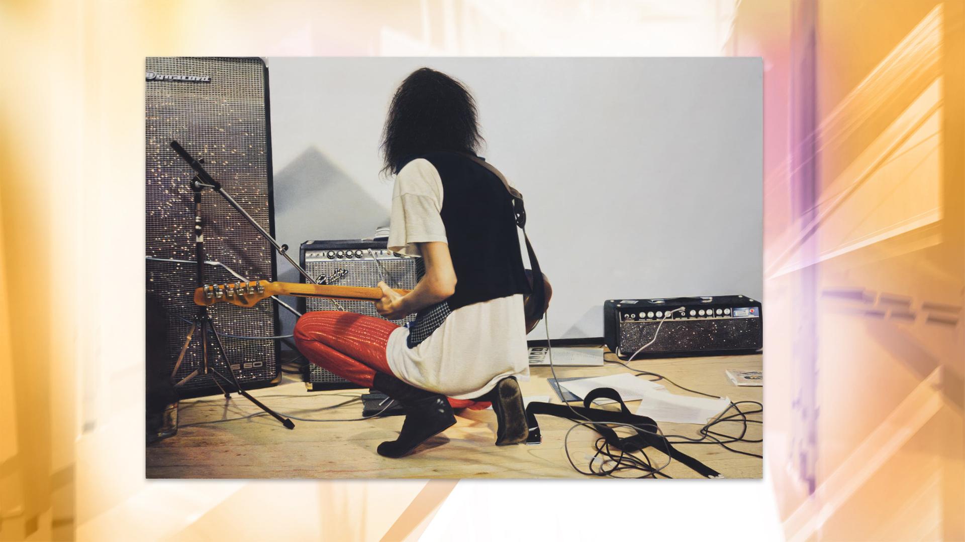 Auf einem fotorealistischen Gemälde sieht man eine Rockmusikerin von hinten, die mit einer Gitarre vor einem Verstärker kniet.
