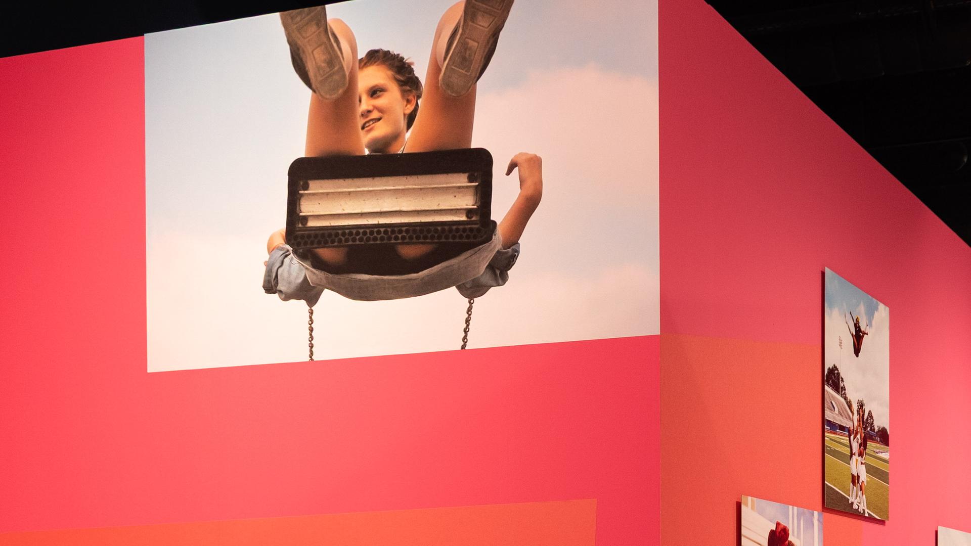 In einem Ausstellungsraum sieht man Fotografien von Menschen an einer roten Wand.