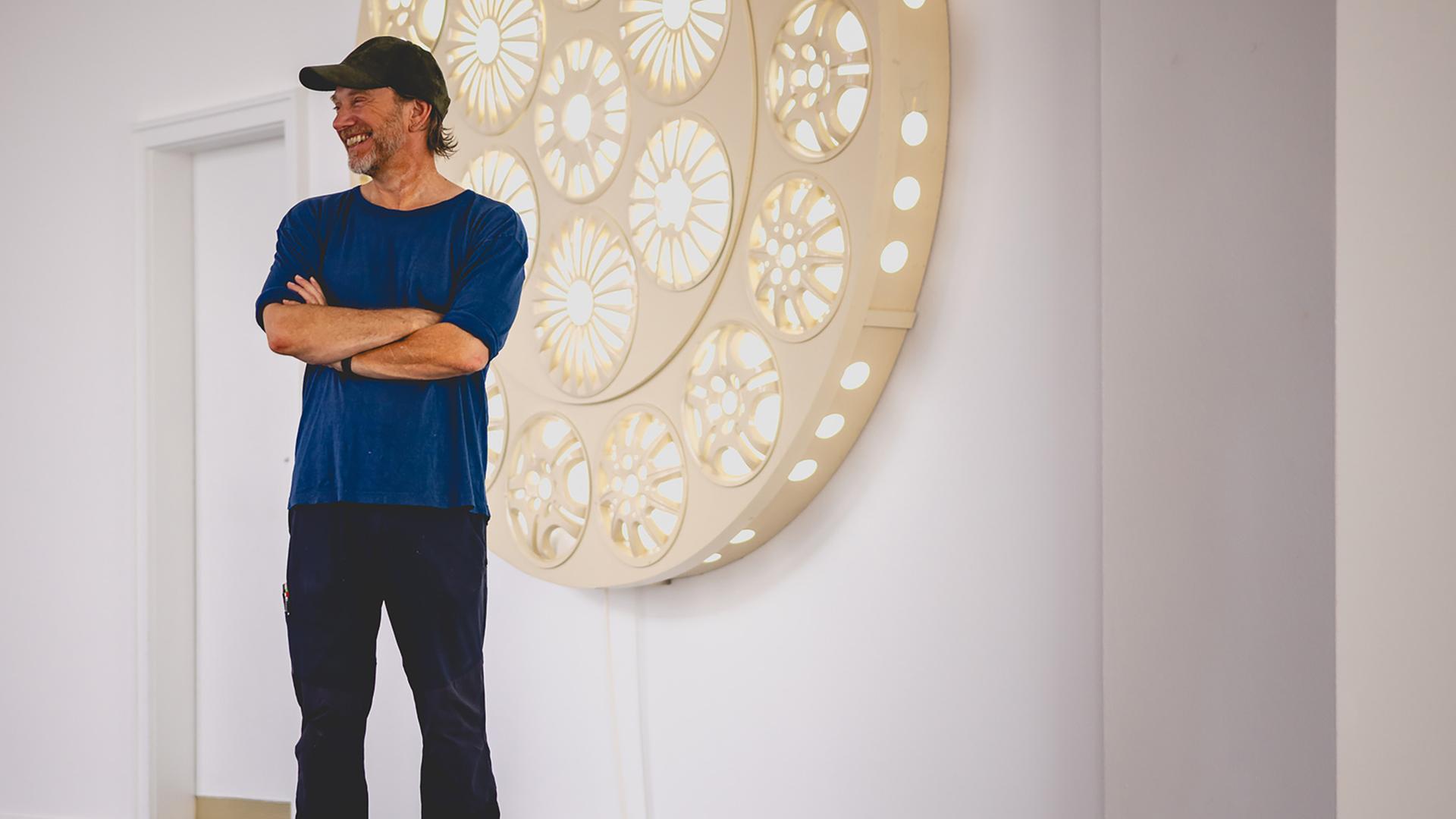 Ein Mann mit Baseballkappe steht vor einem weißen, modernen Kunstwerk, welches an einer Wand hängt.