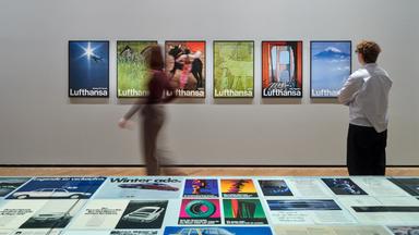 In einem Ausstellungsraum schauen sich zwei Personen bunte Werbefotografien an den Wänden und in den Schaukästen an.