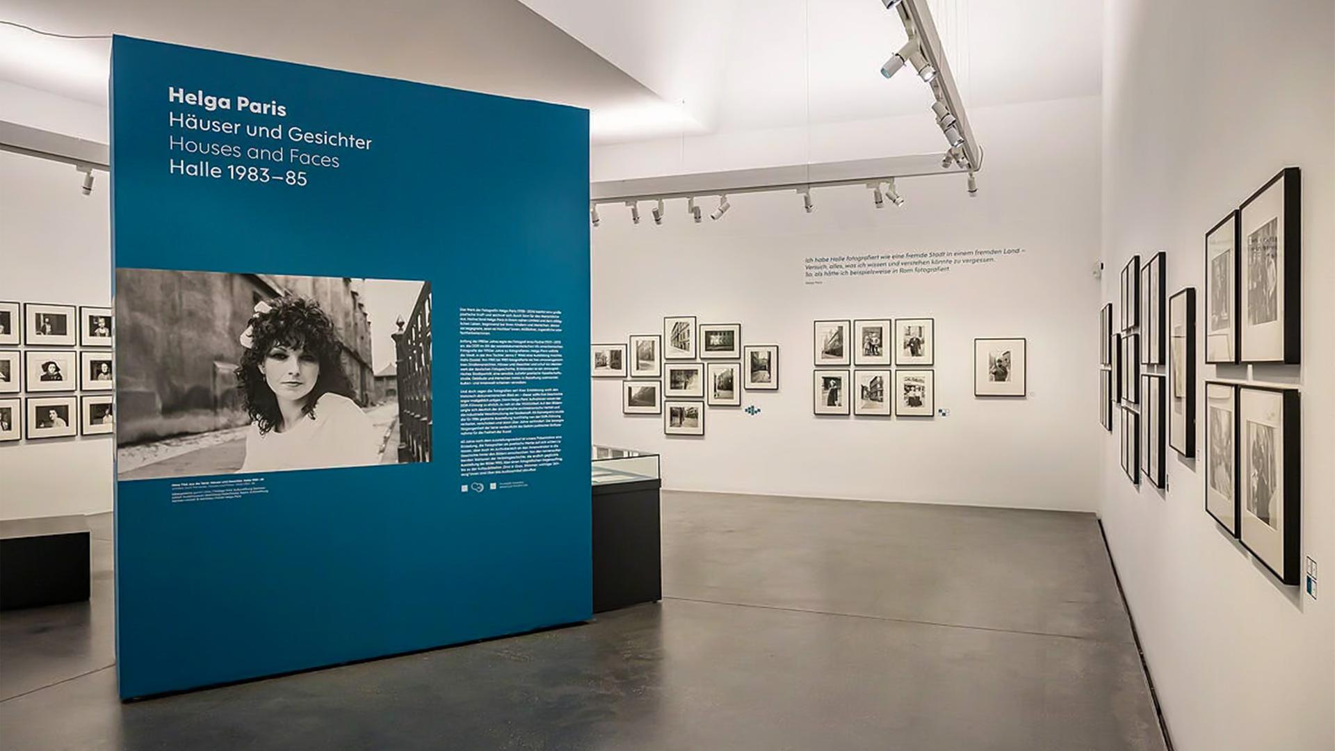 In einem Ausstellungsraum sieht man gerahmte Schwarz-Weiß-Fotografien an den Wänden hängen. Im Raum steht eine groß Stellwand mit einem vergrößerten Foto und Infos dazu.