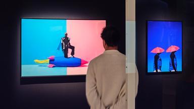 In einem abgedunkelten Ausstellungsraum schaut ein Mann sich ein modernes, buntes Bild an, auf dem eine Person auf einer bunten Rakete steht.