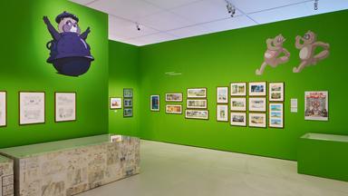 Ausstellungsansicht: Man sieht Bilder an einer grünen Wand hängen und Comic-Zeichnungen von Tieren auf den Wänden.