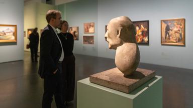 In einem Ausstellungsraum betrachten Menschen an der Wand hängende bunte Gemälde. Im Vordergrund steht die Büste eines Mannes.