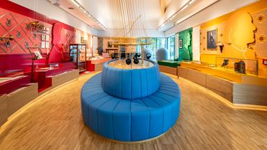 In einem Ausstellungsraum sieht man ein rundes, blaues Sofa umgeben von Infowänden und Ausstellungsstücken.