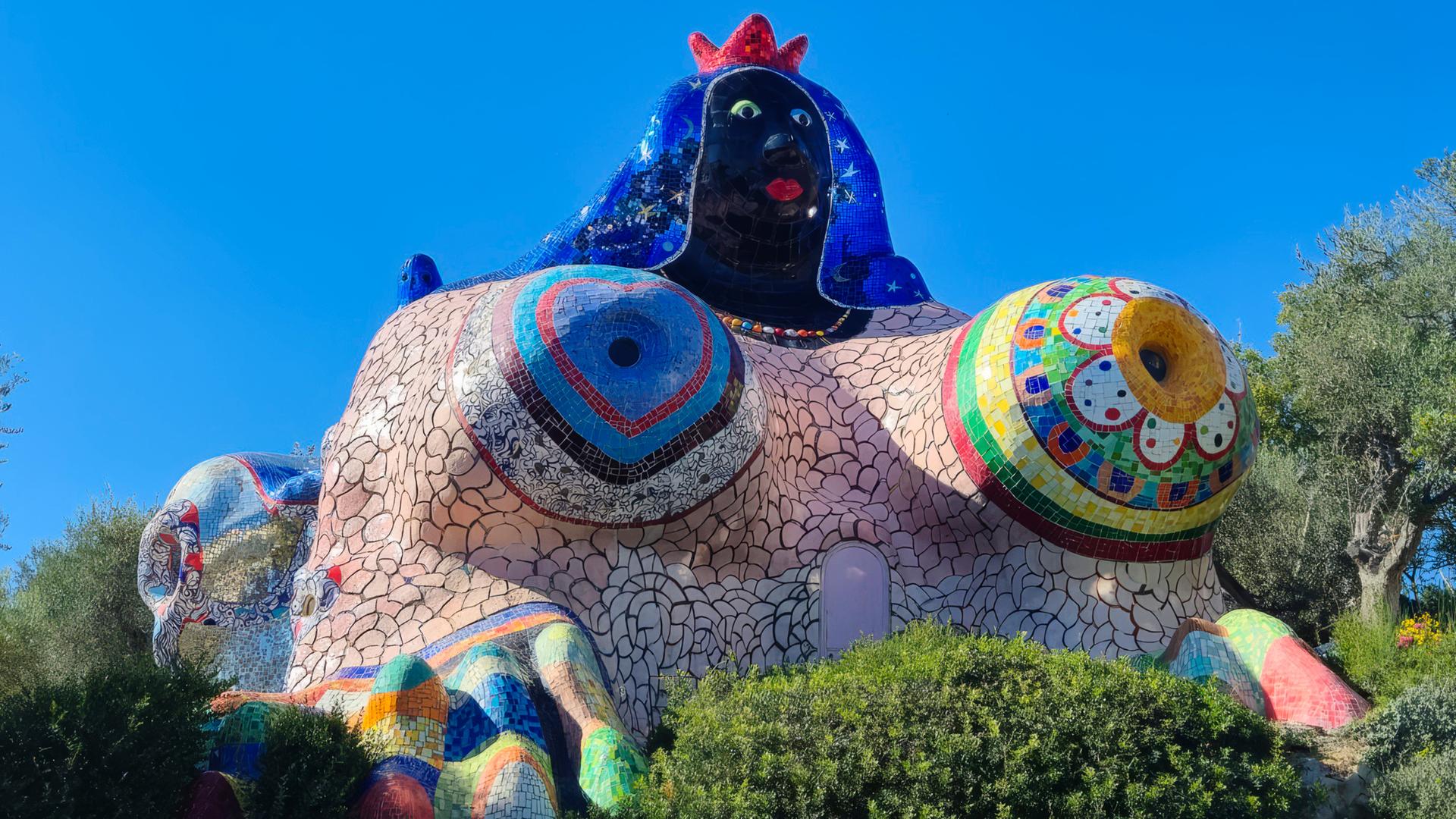 Man sieht ein Gebäude in Form einer voluminösen Frau, das mit buntem Mosaik geschmückt ist.