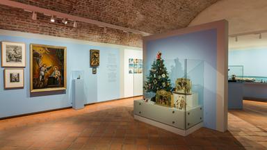 Zu sehen ist ein Raum der Ausstellung. Links an der Wand hängen mehrere Bilder, auf denen Engel zu erkennen sind. Rechts steht ein kleiner geschmückter Weihachtsbaum in einer Vitrine vor einer Wand.