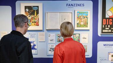 In einem Austellungsraum schauen sich ein Mann und eine Frau Comic-Exponate an.