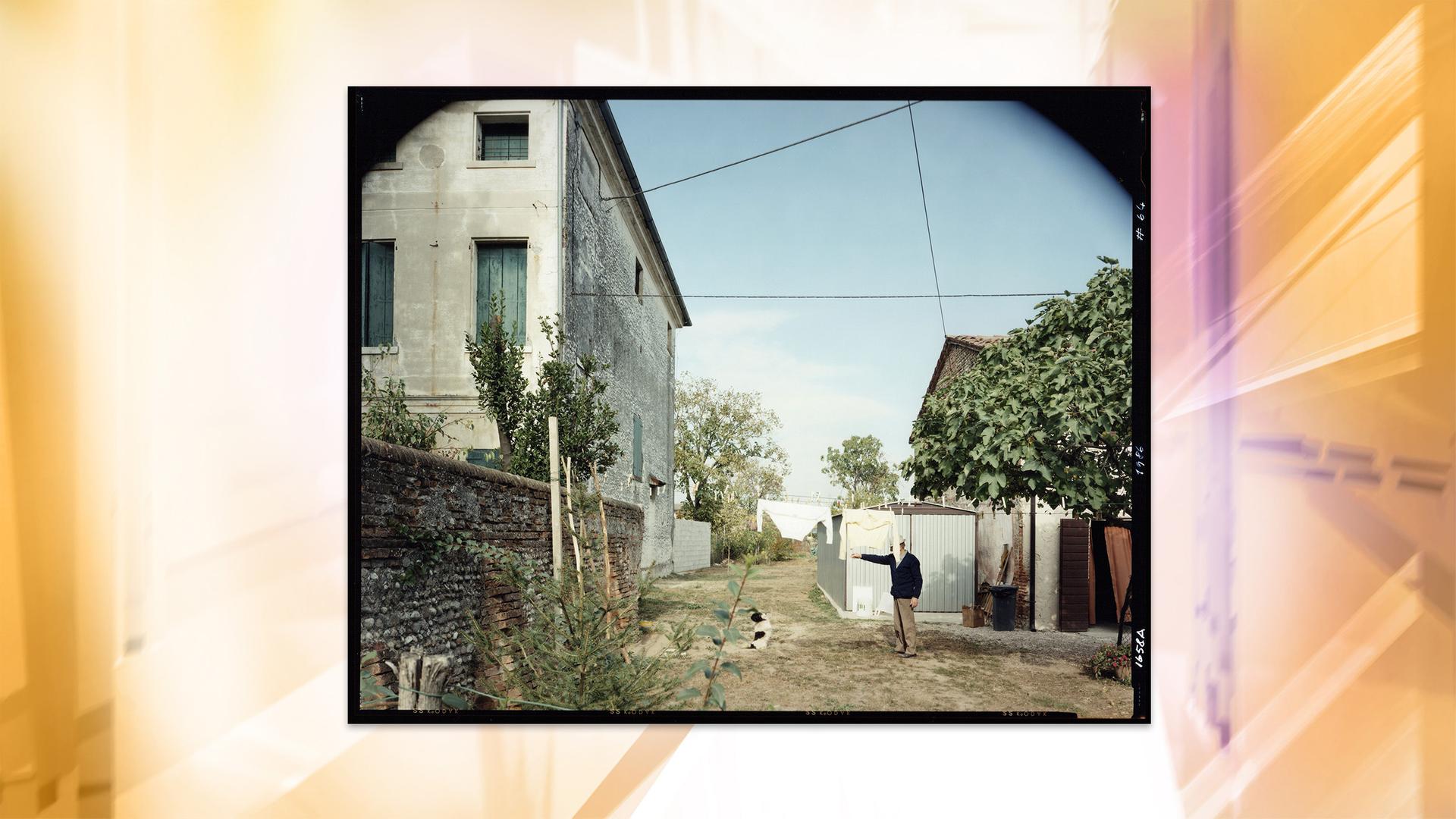 Ein Mann steht in einem Garten zwischen zwei Häusern. Vor ihm sitzt ein Hund, dem er ein Zeichen zu geben scheint. Das Gesicht des Mannes ist von einem Ärmel verdeckt, weil eine Wäscheleine durch den Garten gespannt ist, an der zwei Oberteile hängen. Im Garten stehen Bäume.