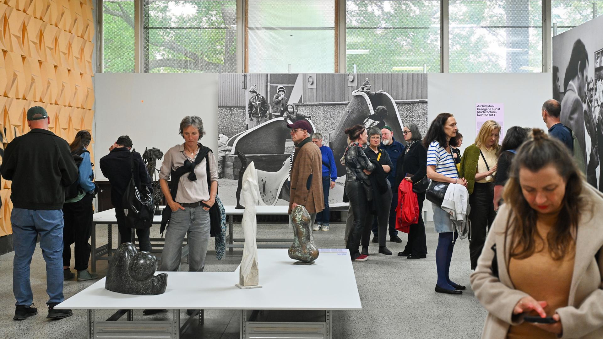 In einem Ausstellungsraum schauen Menschen sich die Plastiken, die auf Tischen stehen und großformatige Schwarz-weiß-Fotografien an den Wänden an.