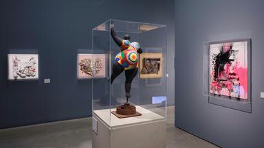 In einem Ausstellungsraum steht eine bunte Frauen-Skulptur in einer Vitrine. An den Wänden hängen moderne Gemälde.