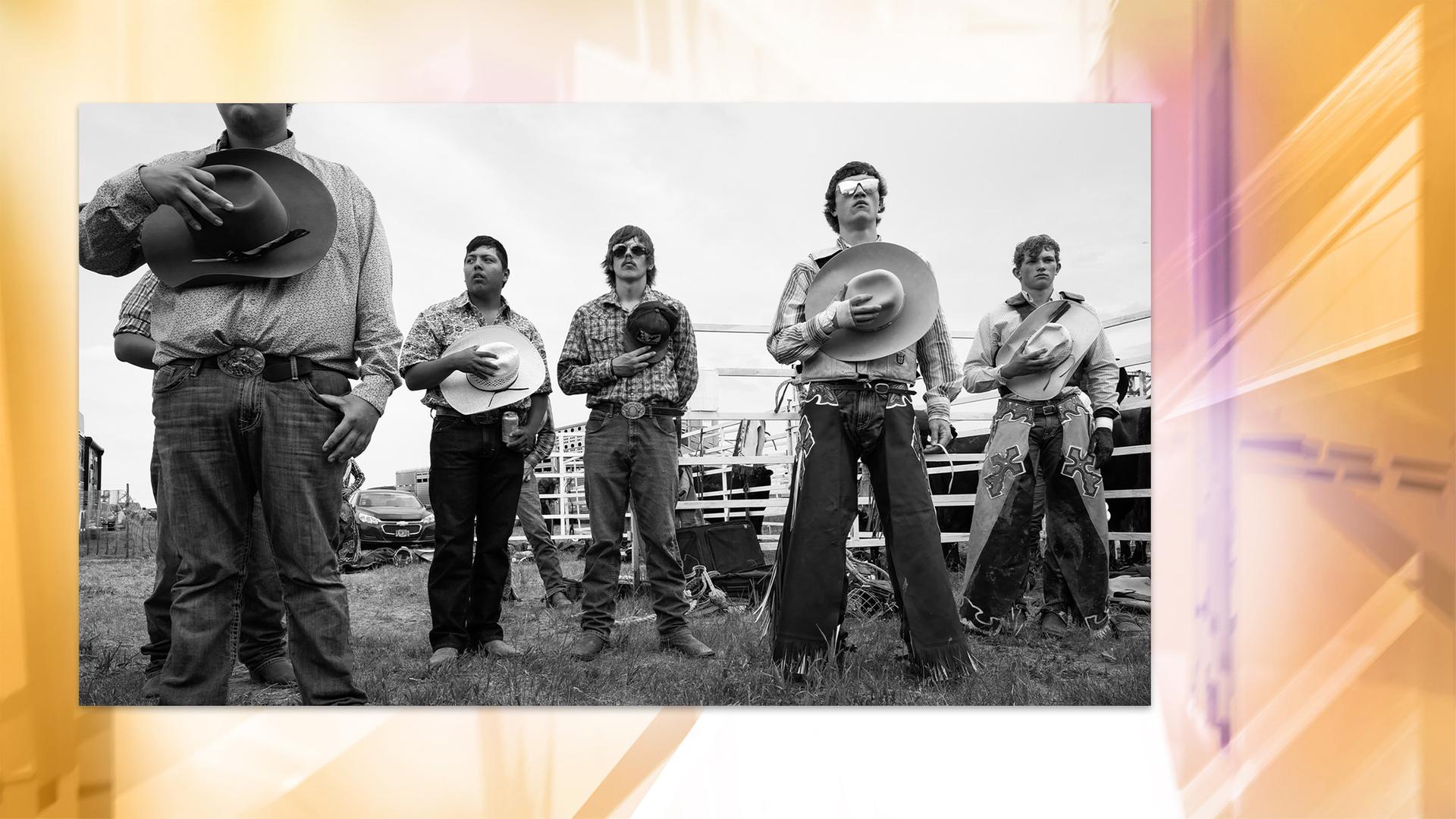 Auf einer Schwarz-Weiß-Fotografie sieht man mehrere breitbeinig nebeneinander stehende Cowboys , die sich jeweils ihren Hut auf die Brust halten.