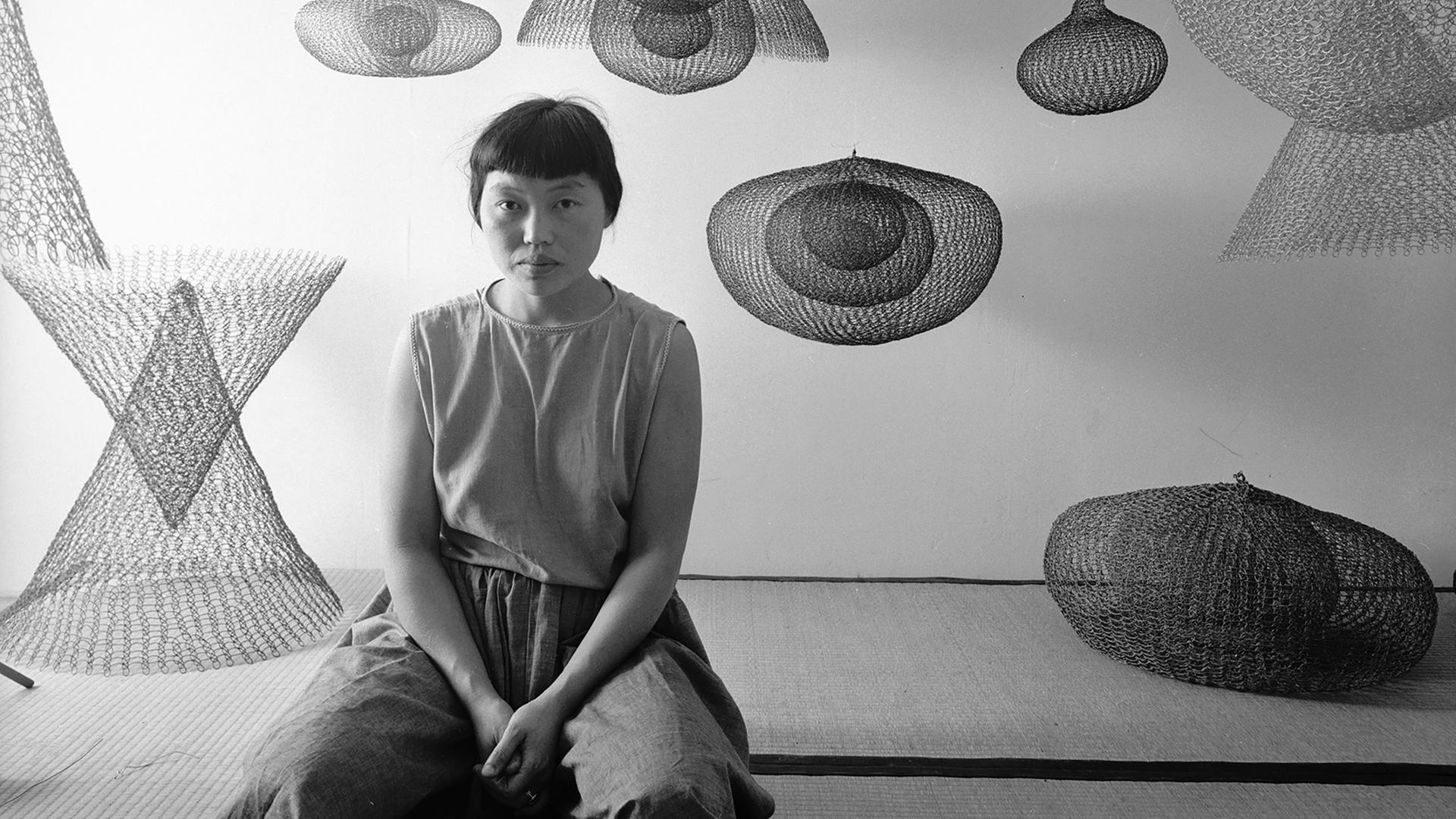 Auf der Schwarz-Weiß-Fotografie sieht man eine kniende Frau, umgeben von Kunstobjekten.
