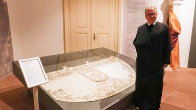 Ein Mann steht neben einer Glasvirtrine, in der ein historischer weißer Umhang mit Goldstickerei liegt.