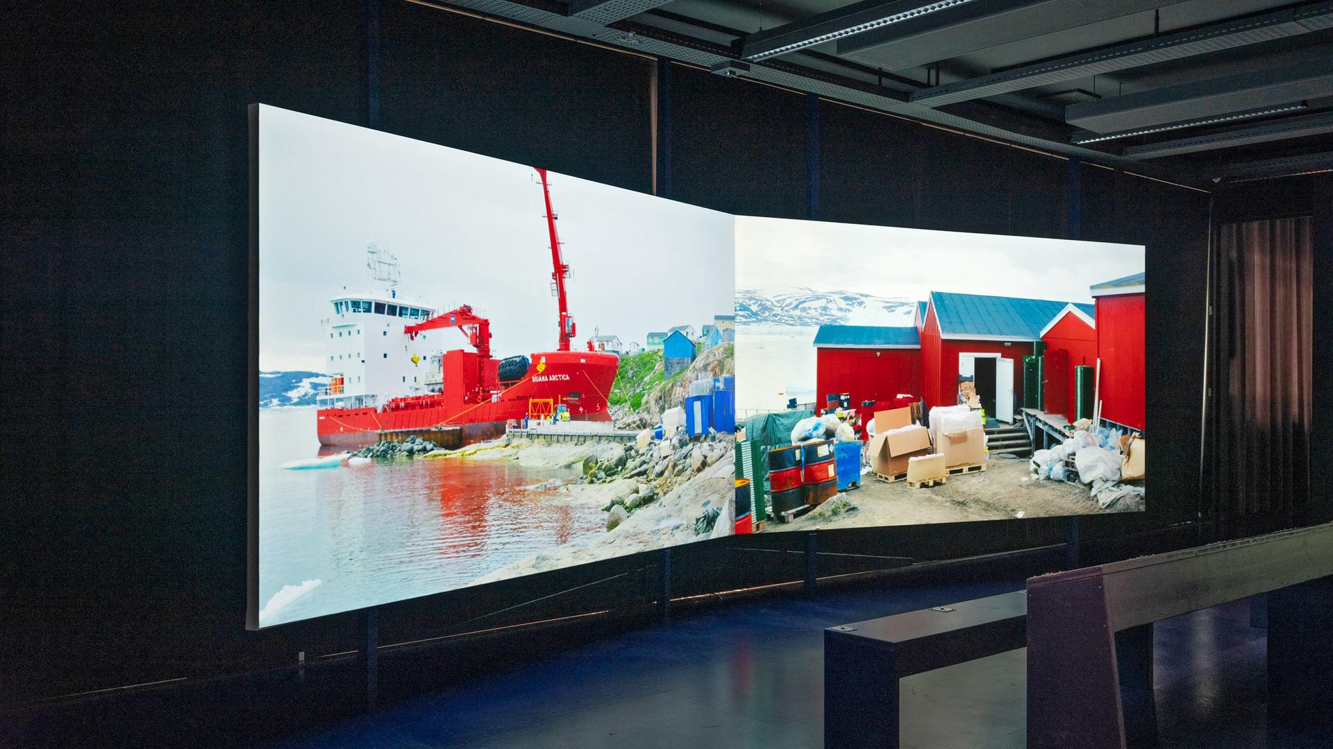 In einem dunklen Raum steht eine zweiteilige Leinwand, auf der ein Schiff an der Küste Grönlands zu sehen ist.