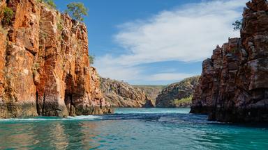 Rötliche, steile Felsen an der Westküste Australiens ragen über türkisfarbenes Meer. Im Hintergrund erstreckt sich die felsige Küstenlandschaft.