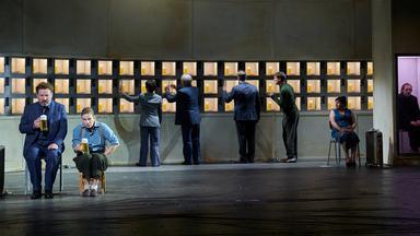 Am linken Bildrand ein freistehender Heizkörper, daneben ein Mann auf einem Stuhl sitzend, er trägt einen dunkelblauen, dreiteiligen Anzug, neben ihm auf einem niedrigen Hocker ein Mann, gespielt von einer Frau, mit hellblauem Hemd, Hosenträgern und dunkelgrüner Hose, beide halten einen vollen Bierkrug in der rechten Hand. Die Rückwand besteht vollständig aus Schließfächern mit gläsernen Türen. Darin befinden sich volle Bierkrüge. Davor befinden sich, mit dem Rücken zum Publikum vier Figuren, die gerade dabei sind, sich Bier zu nehmen. Hinten rechts auf einem Stuhl sitzend eine Frau mit dunkelblauer Kittelschürze, die ihnen dabei zuschaut. In einem abgetrennten Abteil, ähnlich einer Telefonkabine ein weiterer Mann auf einem Stuhl sitzend.