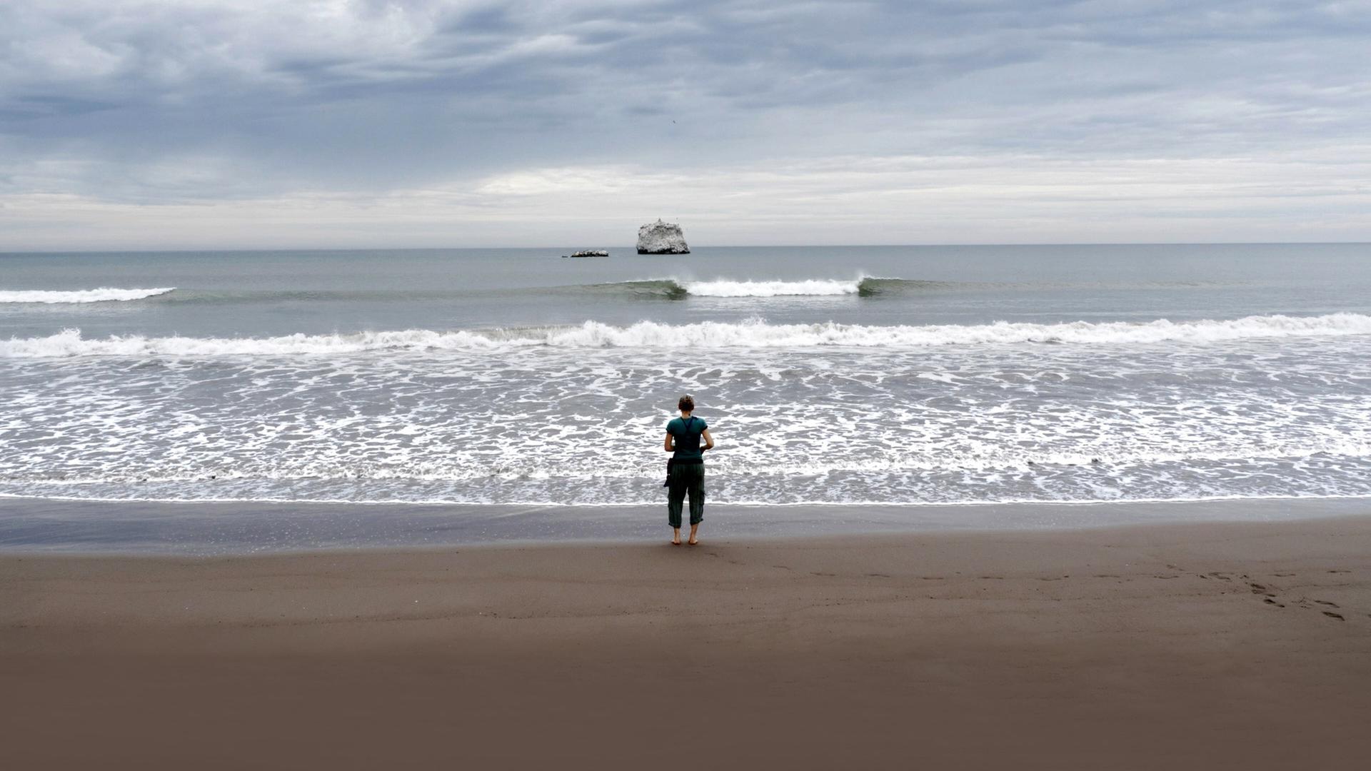 Frau mit dem Rücken zur Kamera am Strand vor Meer.