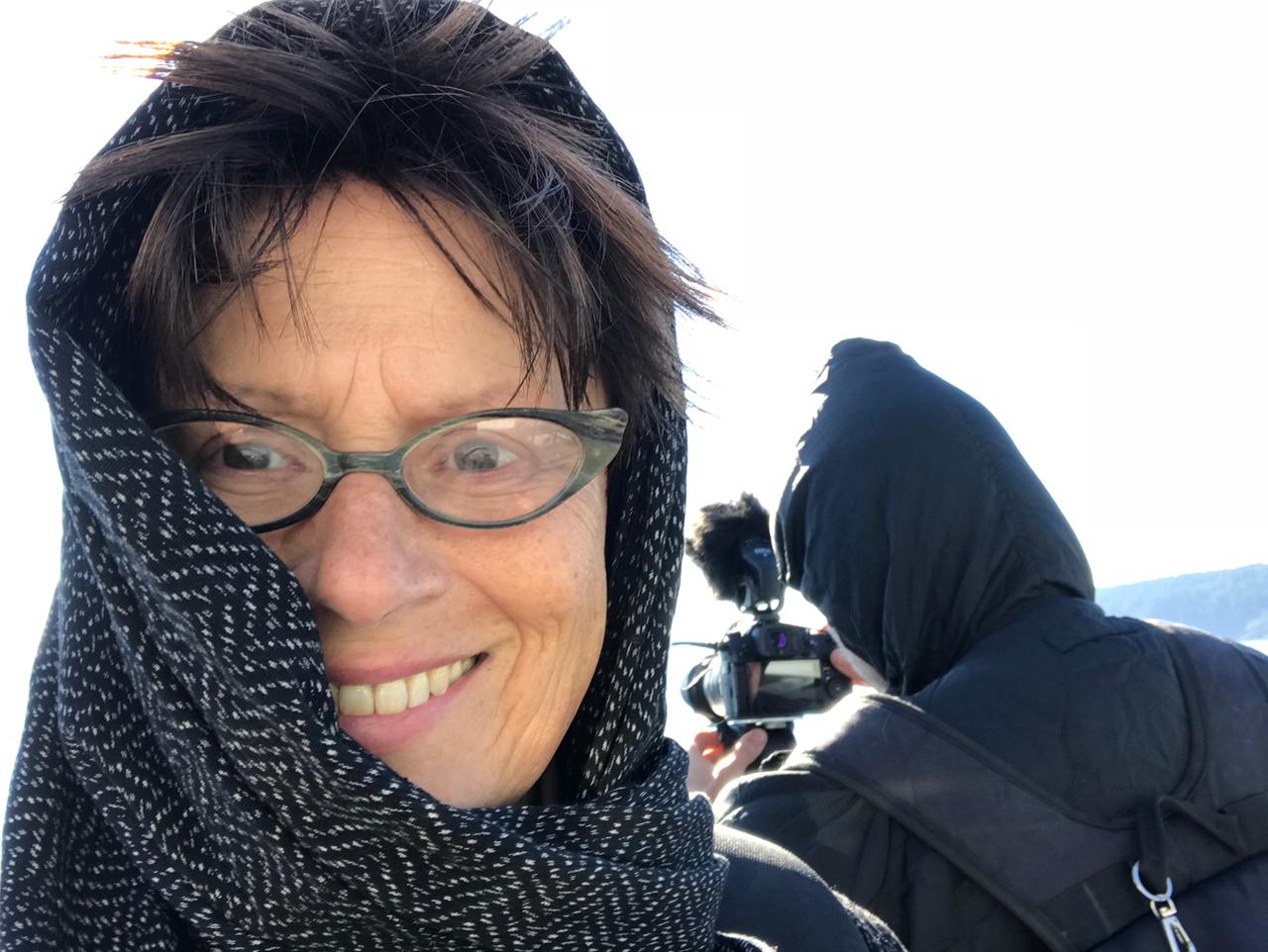 Porträtaufnahme von Filmemacherin Frauke Sandig; sie trägt ein Tuch um Kopf und Hals, das teils ihr Gesicht verdeckt; sie hat eine modische Brille auf und lächelt; im Hintergrund ist ein Kameramann von hinten zu sehen, der einen Anorak trägt und sich die Kapuze übergezogen hat.
