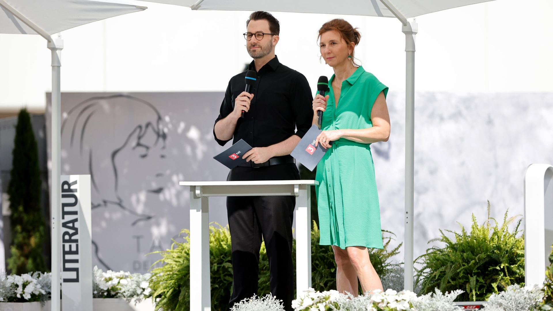 Zwei Moderator:innen stehen unter weißen Sonnenschirmen auf einer Bühne im Freien und sprechen in Mikrofone; im Hintergrund ist das Logo der Tage der deutschsprachigen Literatur sichtbar.
