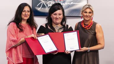 Drei Frauen stehen nebeneinander und präsentieren glücklich lächelnd zwei rote Kladden mit zwei Preisurkunden.