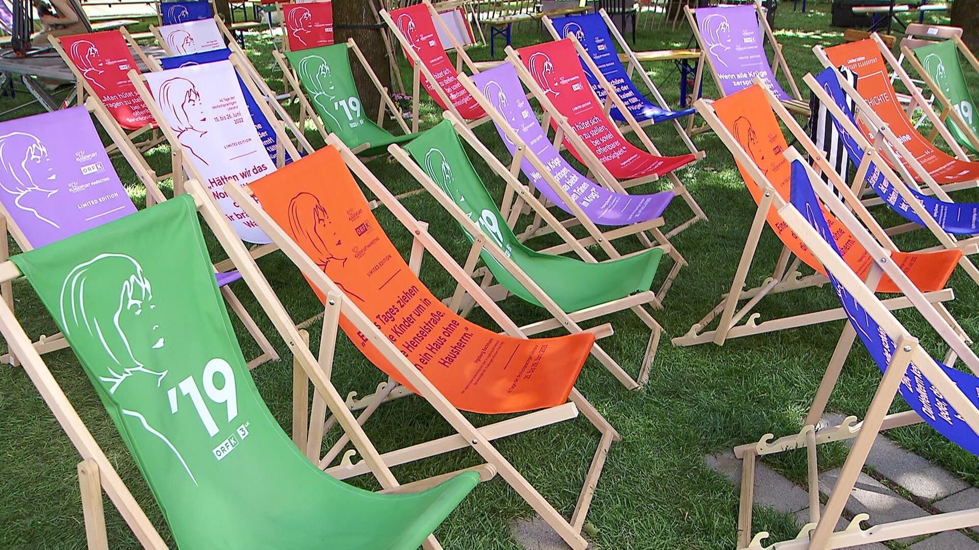 Das Bild zeigt eine Anordnung von Liegestühlen, die in einem Park aufgestellt sind. Die Stühle sind in verschiedenen Farben gestaltet, darunter Grün, Orange, Lila, Blau und Rot. Auf den Stühlen sind unterschiedliche Texte und Silhouetten abgebildet, die auf literarische Themen und den Ingeborg Bachmann-Preis hinweisen. Der Boden ist mit Rasen bewachsen, und die Stühle sind in lockerer Reihenfolge aufgestellt, so dass zwischen ihnen genügend Platz ist. Im Hintergrund sind weitere Strukturen oder Möbel zu erahnen, wobei die Atmosphäre entspannt wirkt, passend zu einer Lesung oder einer Literaturveranstaltung.