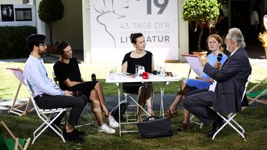 Vier Personen werden von einem Mann interviewt. Sie sitzen um einen Tisch im Freien.