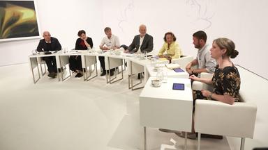 Sieben Personen sitzen im Studio an Tischen in einem Halbrund.