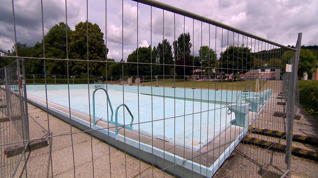 Das Bild zeigt ein verlassenes Freibad, das durch ein Metallgitter eingezäunt ist. Im Vordergrund ist der leere Schwimmbecken sichtbar, dessen Wände in einem hellen Blau gehalten sind. Eine Schwimmleiter steht auf der linken Seite des Beckens.   Im Hintergrund sind große, grüne Bäume zu sehen, die einen Teil der Umgebung umrahmen. An einer Stelle sind einige Gebäude erkennbar, wahrscheinlich Gemeinschaftseinrichtungen oder Umkleideräume des Freibades. Der Himmel ist bewölkt, was auf eine mögliche Wetteränderung hinweist und der Szene einen melancholischen Eindruck verleiht.   Dies ist ein Ort, der anscheinend in Vergessenheit geraten ist, was durch die Abwesenheit von Menschen und das eingezäunte Becken betont wird. Am Rande des Feldes sind einige Bänke und einfache Einrichtungen sichtbar. Der Gesamtzustand des Freibades deutet auf einen Mangel an Pflege und die Herausforderungen hin, vor denen öffentliche Schwimmbäder stehen.