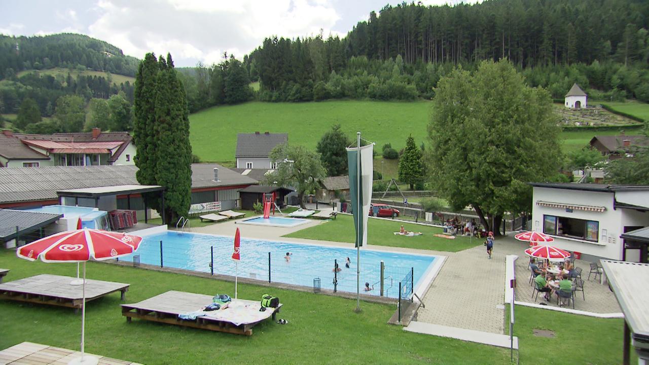 Das Bild zeigt ein Freibad in einer ländlichen Umgebung, wahrscheinlich in Österreich. Im Vordergrund befindet sich ein rechteckiger Swimmingpool, in dem einige Schwimmer zu sehen sind. Um den Pool herum stehen mehrere Liegen und Sonnenschirme, darunter rote mit weißen Streifen. Auf der rechten Seite des Pools gibt es ein Gebäude mit einem überdachten Bereich, wo Menschen an Tischen sitzen. Im Hintergrund sind grüne Wiesen und Bäume zu sehen, die die ländliche Kulisse ergänzen. Einige Häuser sind ebenfalls in der Nähe des Schwimmbads sichtbar, wobei die Dächer einige unterschiedliche Bauweisen aufweisen. Hinter den Gebäuden erhebt sich ein bewaldeter Hang. Die Atmosphäre wirkt entspannt und einladend.