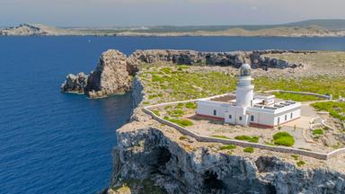 Ein weißer Leuchtturm mit Nebengebäuden steht auf einer hohen, steilen Kalksteinklippe am Cap Cavalleria, umgeben von grünem Gestrüpp und mit Blick auf das tiefblaue Meer.