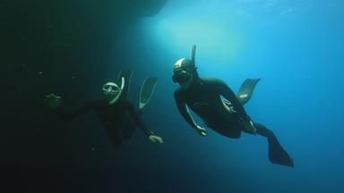 Zwei Apnoe-Taucherinnen in Neoprenanzügen mit Schnorcheln und Flossen tauchen unter Wasser vor der felsigen Küste Ibizas.