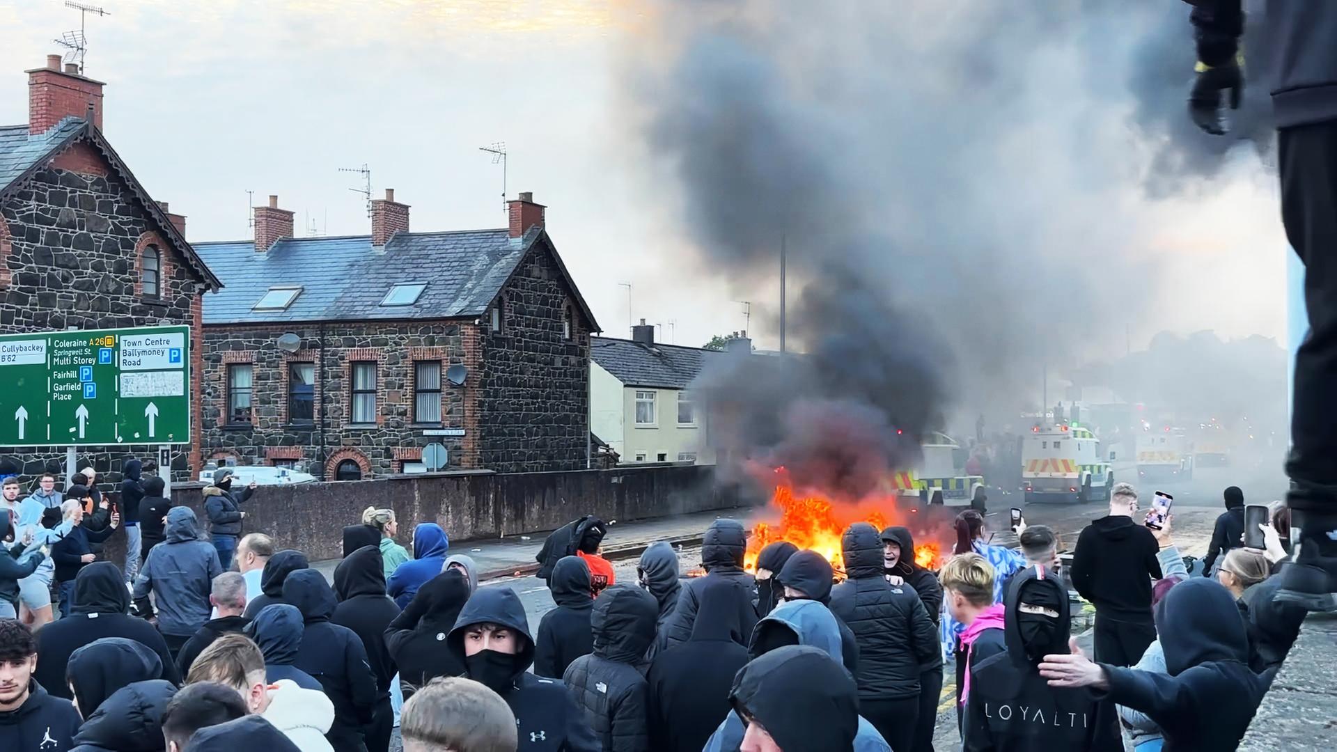 Eine große Gruppe vermummter Menschen steht auf einer Straße, auf der etwas brennt. Hinter dem Feuer sind Einsatzfahrzeuge zu sehen.