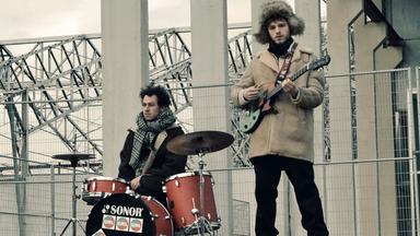 Ein junger Mann mit Gitarre und einer mit einem Schlagzeug stehen vor einer Brückenkonstruktion aus Beton und Stahl.