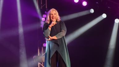 Das Bild zeigt eine weibliche Schauspielerin auf einer Bühne. Sie hat schulterlange, lockige blonde Haare und trägt ein langärmliges Oberteil in dunklem Blau mit helleren Akzenten sowie eine schwarze Hose. Die Künstlerin hat ein freundliches, einladendes Lächeln und gestikuliert, während sie sich dem Publikum zuwendet.  Im Hintergrund ist eine Bühnenbeleuchtung zu sehen, die in verschiedenen Farben leuchtet, insbesondere in Purpur und Weiß. Die Lichter werfen Strahlen auf die Bühne und erzeugen eine dynamische Atmosphäre. Rechts von ihr steht ein schwarzer Stuhl, und auf der Bühne sind schwache Nebelschwaden zu erkennen.   Die gesamte Szenerie vermittelt eine lebhafte, unterhaltsame Stimmung, die auf eine Performance hindeutet.