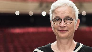 Eine Frau mit grauen Haaren, Brille und roten Lippen lächelt dezent in die Kamera. 