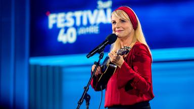 Barbara Ruscher steht auf der rechten Seite des Bildes. Sie hält eine Ukulele in der Hand. Sie lächelt und trägt ein rotes Haarband.