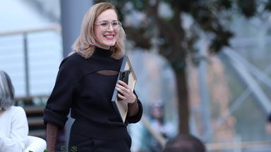 Eine junge blonde Frau mit Brille trägt lächelnd ein gerahmtes Dokument unter dem Arm und läuft durch einen Raum.