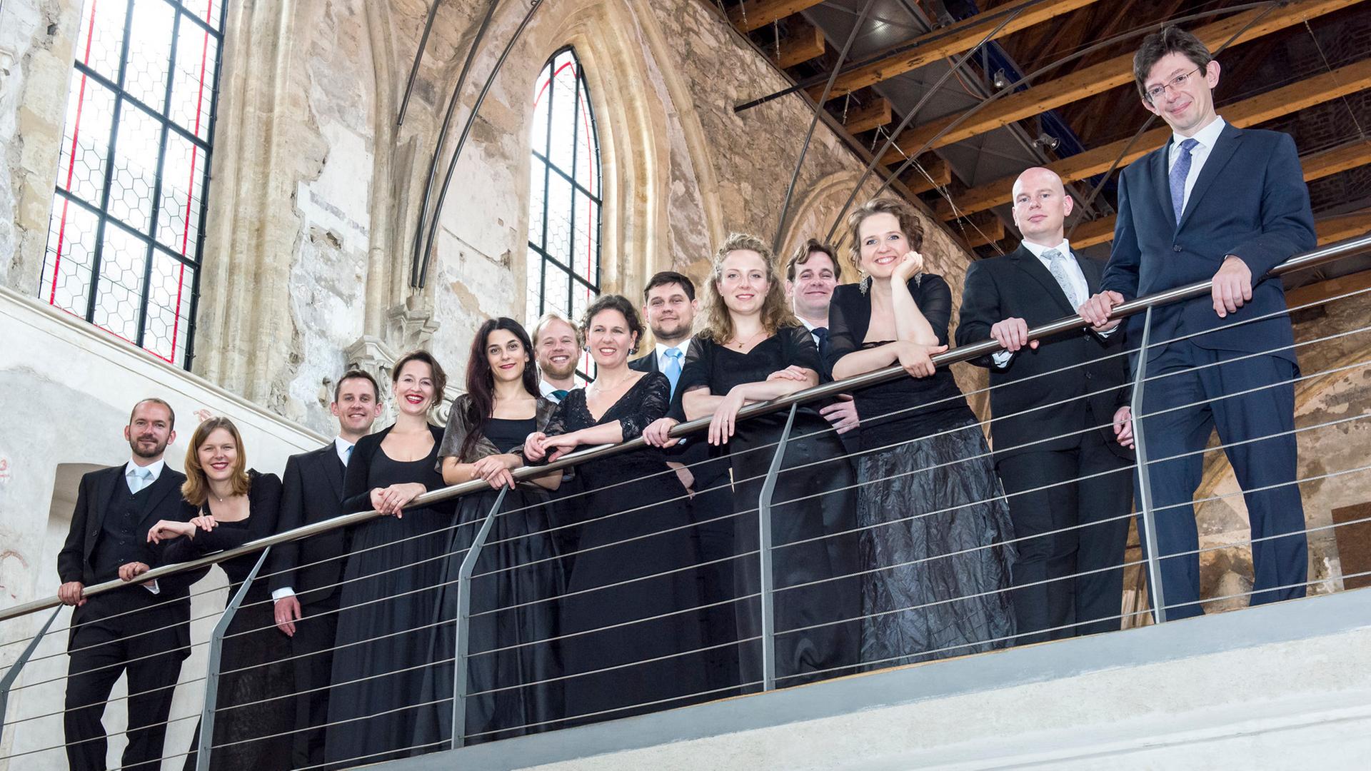 Gruppenfoto des Vokalensembles Collegium Vocale, das in eleganter Abendgarderobe auf einer Balustrade in einem historischen Kirchengebäude steht. Die Mitglieder lächeln in die Kamera, hinter ihnen hohe gotische Fenster und eine Holzdecke.