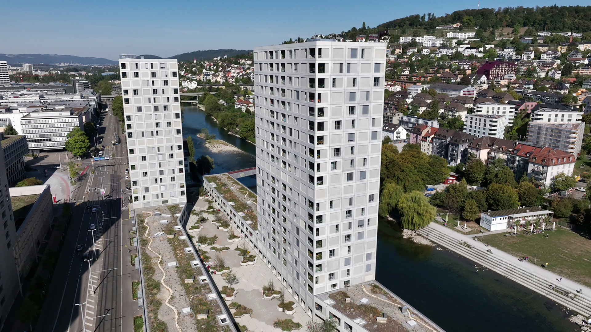 Das Bild zeigt zwei hohe, moderne Wohngebäude mit einer strukturierten Fassade. Im Vordergrund verläuft eine Straße, während im Hintergrund Bäume und weitere Gebäude zu sehen sind. Ein Fluss fließt neben den Gebäuden.