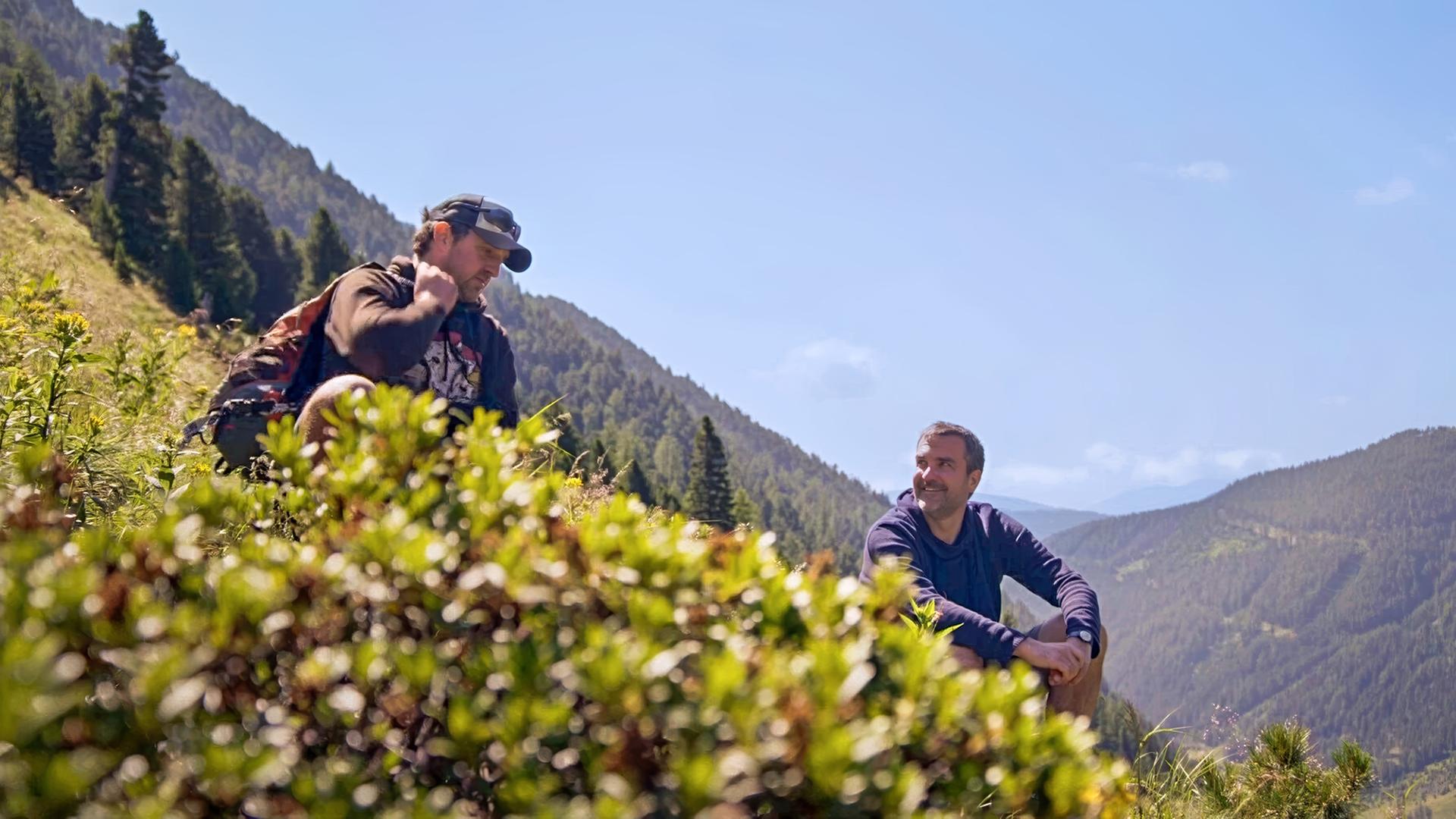 Zwei Männer sitzen auf einer Almwiese inmitten der Berge und unterhalten sich  einer mit Rucksack und Kappe, der andere lächelnd, mit Blick auf seinen Gesprächspartner; umgeben von sommerlicher Vegetation und Nadelwald.