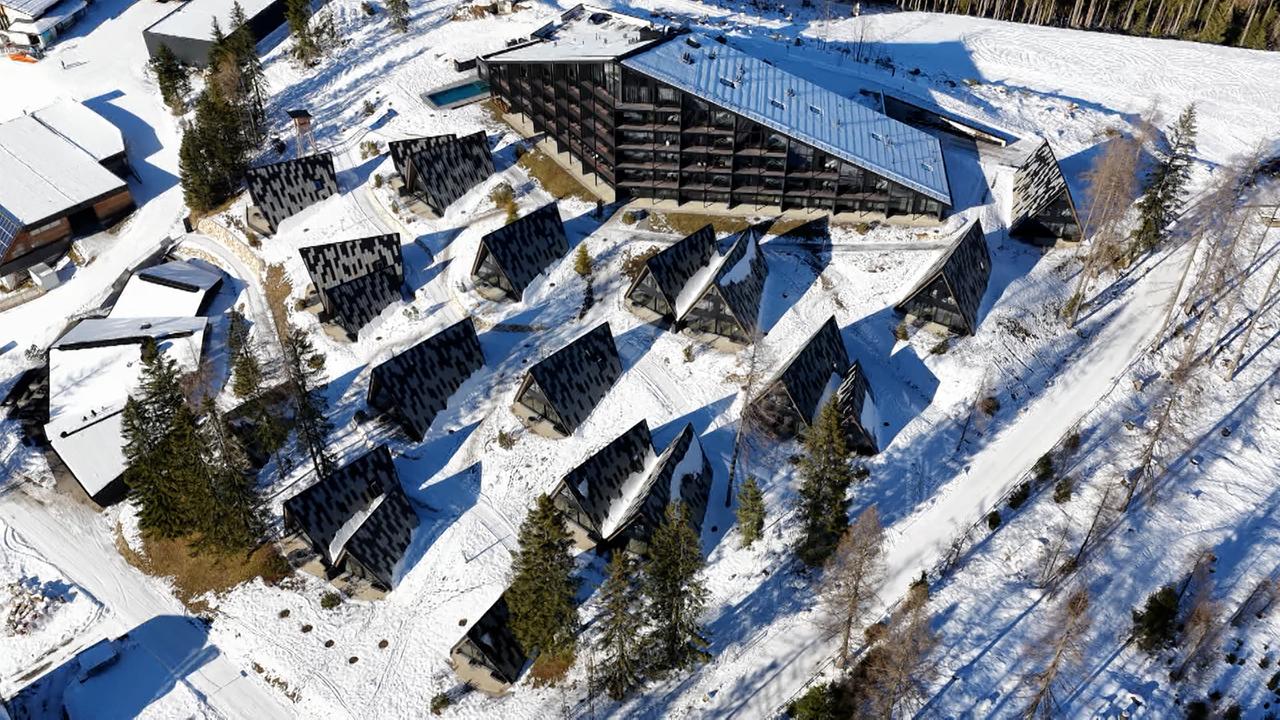 Das Bild zeigt eine Luftaufnahme einer Hotelanlage, die von Schnee umgeben ist. Im Zentrum steht ein großes, modernes Gebäude mit einem flachen Dach, das in dunklen Farbtönen gehalten ist und breite Fenster hat. Um dieses zentrale Gebäude sind mehrere kleinere, chaletartige Strukturen angeordnet, die ebenfalls ein schräges Dach besitzen. Diese sind in einem ähnlichen, dunklen Farbton gestaltet, und einige Dächer sind mit Schnee bedeckt.  Der gesamte Bereich ist von Nadelbäumen umgeben, die an den Rändern des Grundstücks stehen. Wege sind im Schnee sichtbar und führen zu den verschiedenen Gebäuden. Das Bild vermittelt den Eindruck einer ruhigen, alpinen Umgebung, die sowohl zur Erholung als auch für winterliche Aktivitäten genutzt werden könnte.